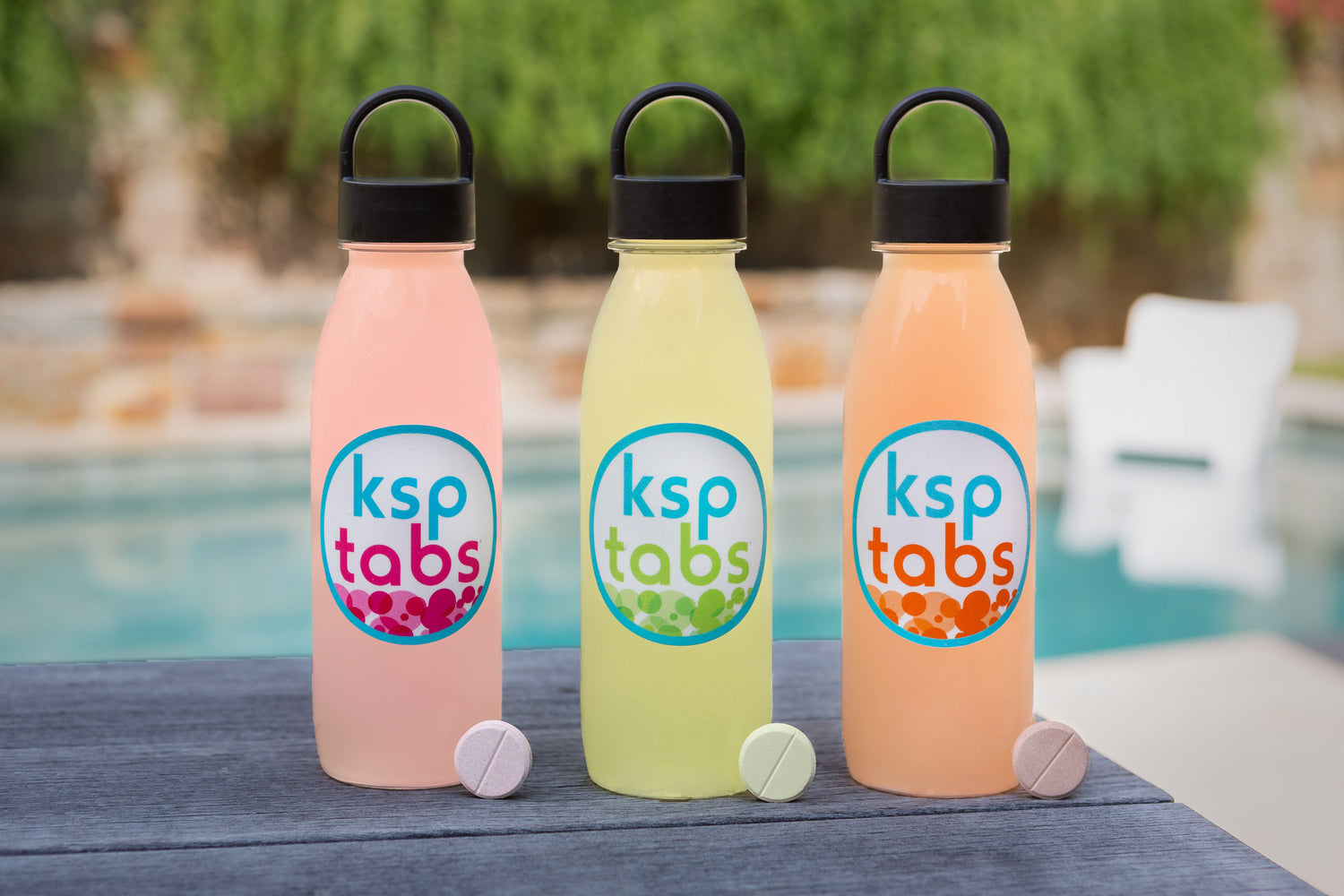 Three colorful water bottles with 'ksp tabs' labels on a table by a pool.