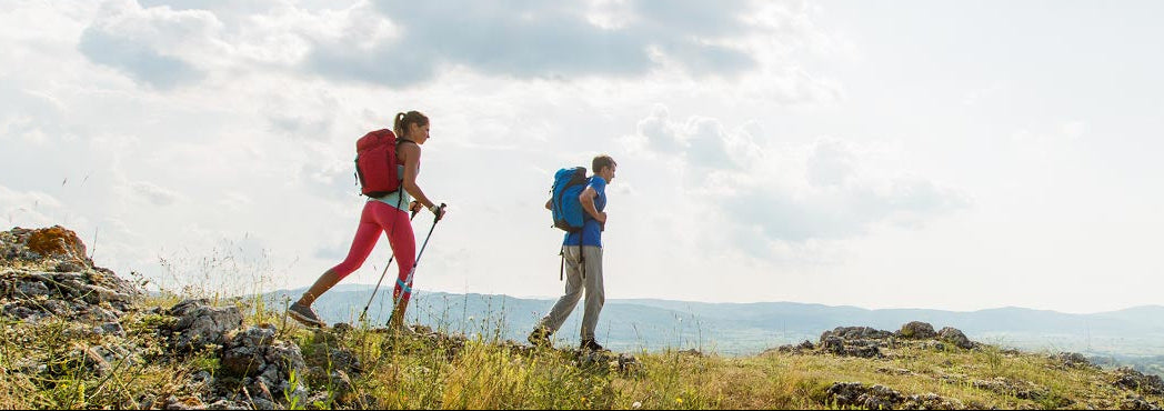 Two hikers with backpacks walking on a rocky trail with a scenic background.